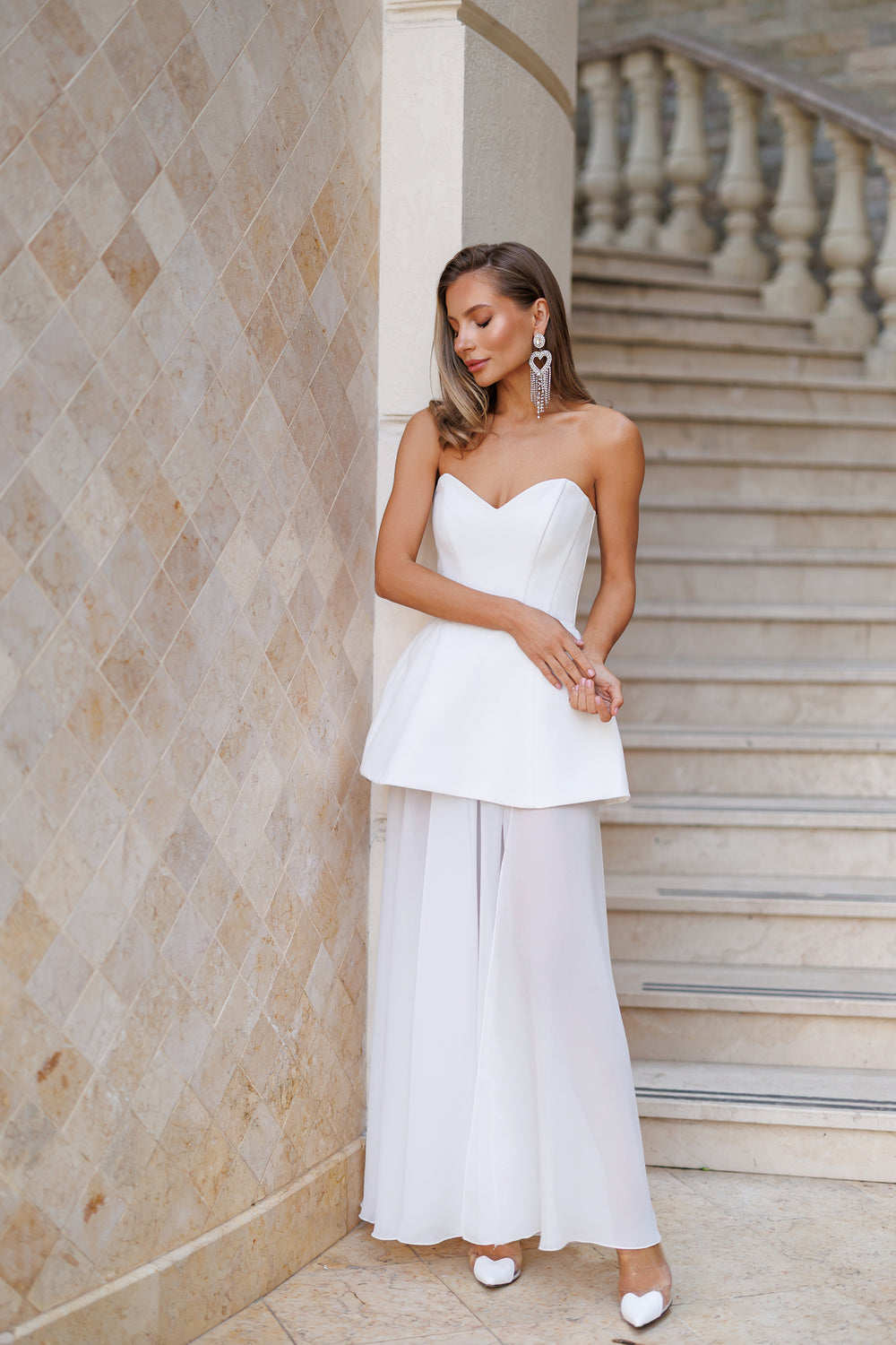 Woman in a white strapless top and wide-leg pants standing against a stone wall with steps.