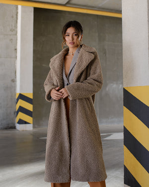 Woman wearing a brown teddy coat in an industrial setting with concrete walls and yellow and black striped barriers.