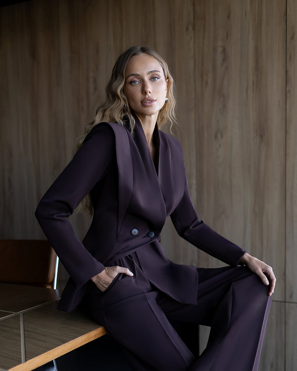 Woman in a dark purple suit sitting against a wooden wall.