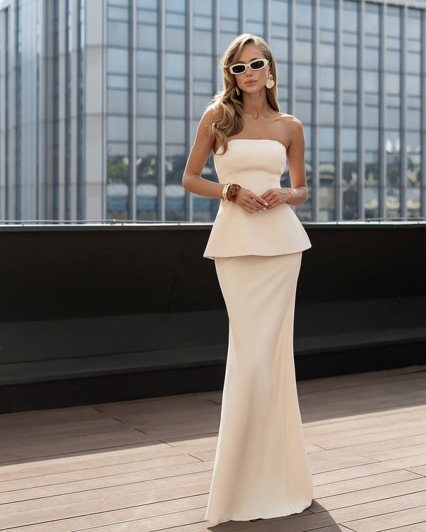 Woman in a strapless white peplum dress standing on a rooftop with a cityscape background.
